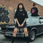 A scallywag sportin' a black oversized tee with an embroidered chest logo and bold back graphic, set against a vintage car and graffiti.