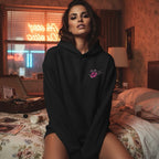 Black hoodie with embroidered logo, oversized fit, model sittin' on a bed with neon sign and vintage suitcase in the background.