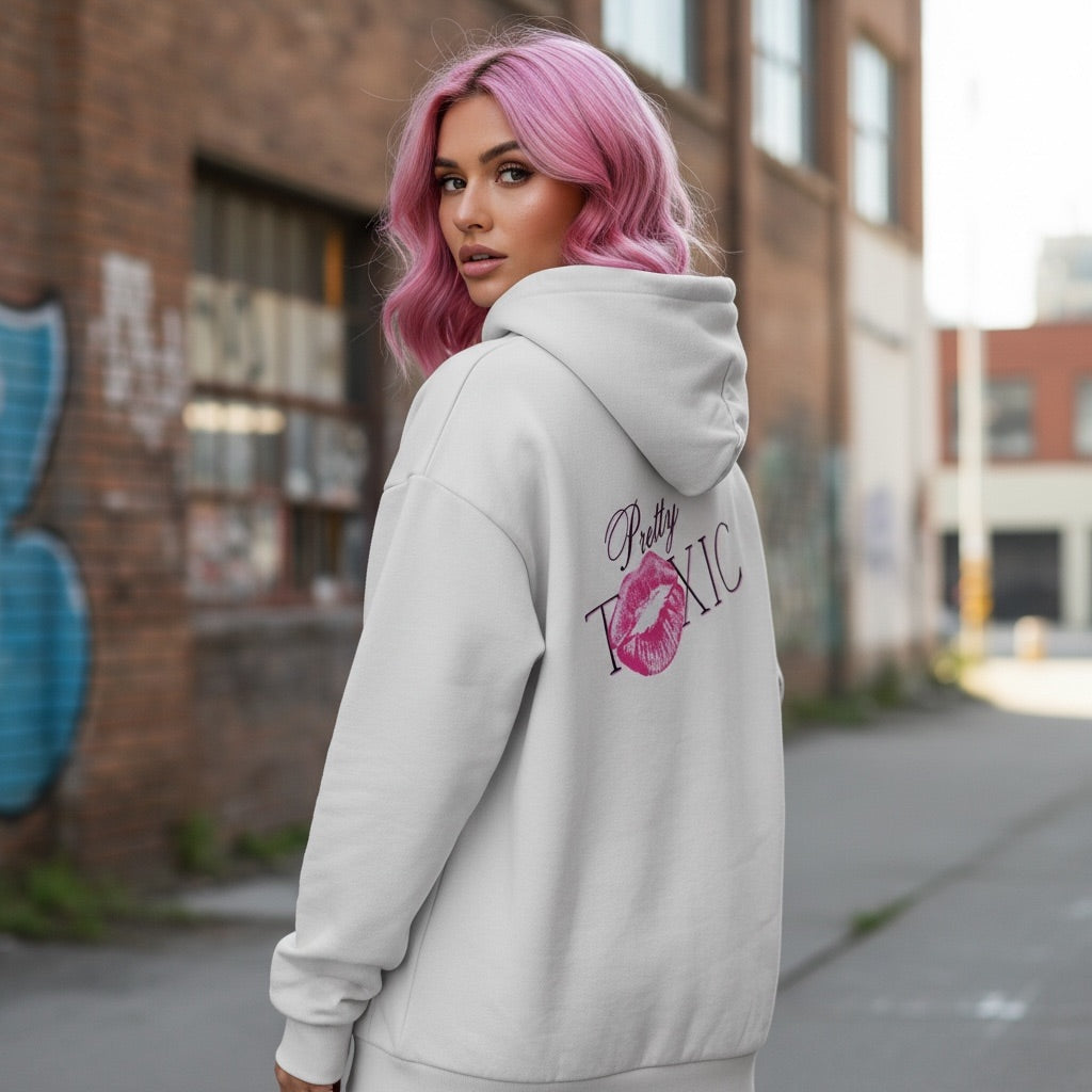 Light-colored oversized hoodie with 'Pretty Toxic' graphic and kiss mark, worn by a model with pink hair in an urban setting.