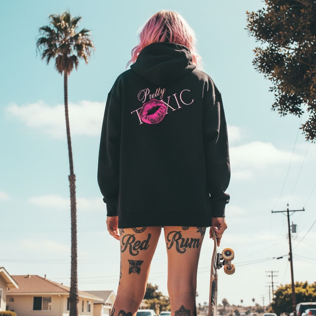 Black oversized hoodie with bold 'Pretty Toxic' graphic and lip print, model holdin' skateboard by palm trees.